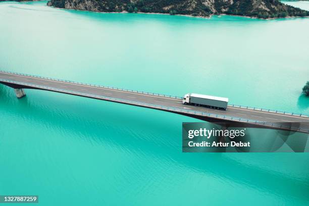 drone view following semi truck driving on bridge crossing blue lake in the pyrenees mountains. - truck birds eye stockfoto's en -beelden