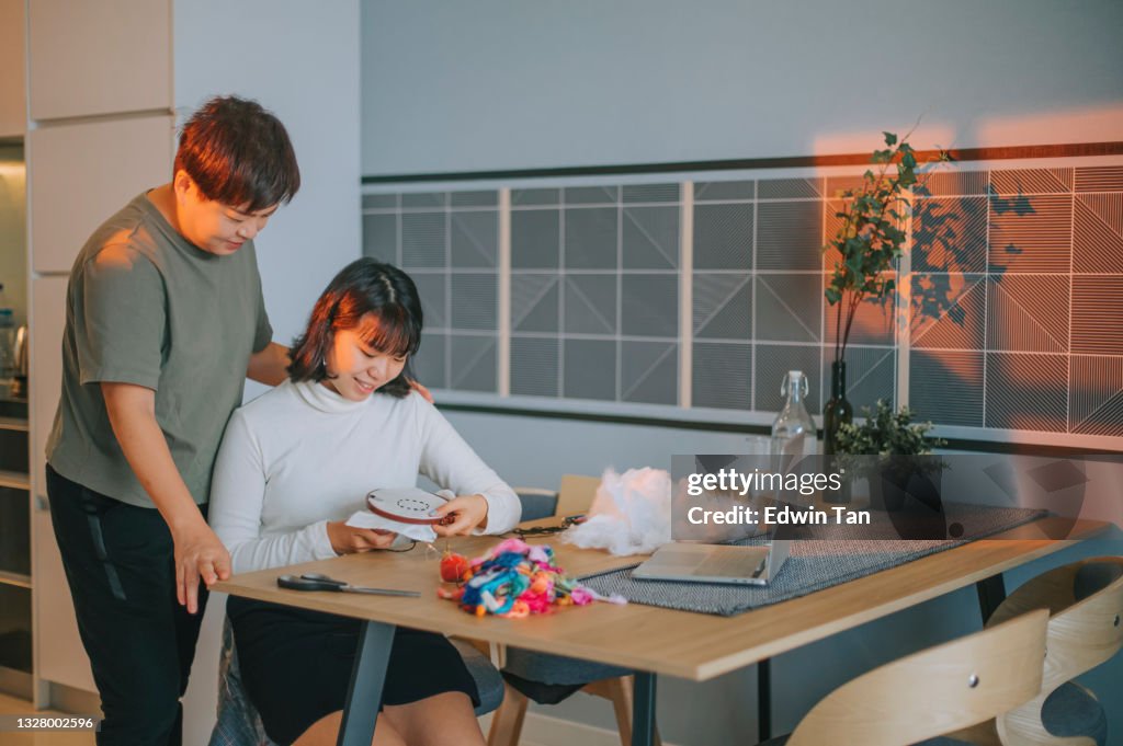 Adolescente china asiática y madre Punto de cruz bordado hecho a mano durante la noche aprendiendo de Internet con la computadora portátil en el ajetreo lateral de casa