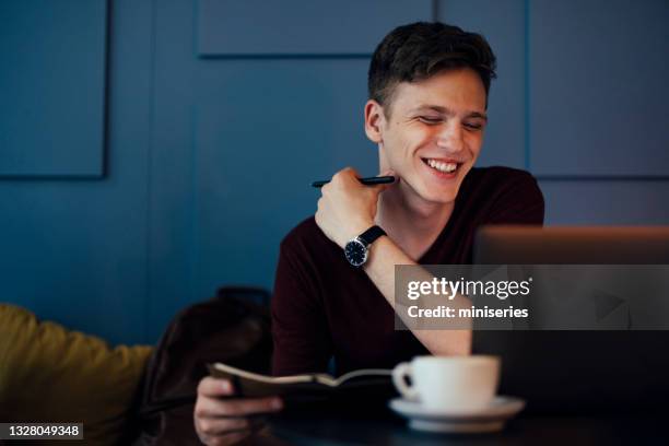 studente maschio allegro che studia sul computer portatile mentre beve caffè in una caffetteria - notepad white table foto e immagini stock