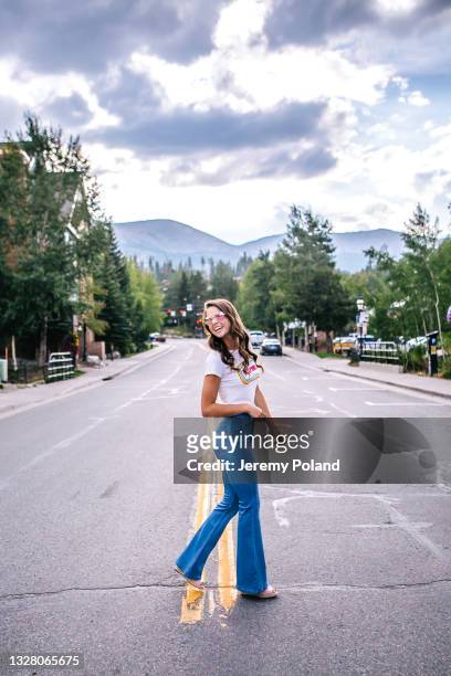 ritratto a figura intera di una bella e alla moda giovane donna che indossa jeans flare e occhiali da sole che attraversa la strada in una città di montagna del colorado - pantaloni a zampa di elefante foto e immagini stock