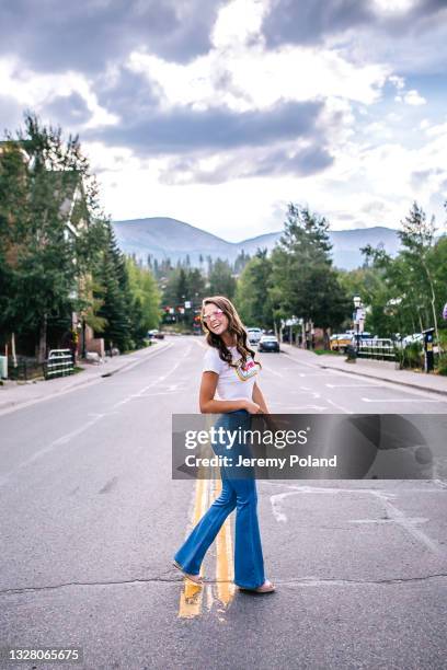 full length portrait of a beautiful, fashionable young woman wearing flare jeans and sunglasses walking across the street in a colorado mountain town - flared trousers imagens e fotografias de stock