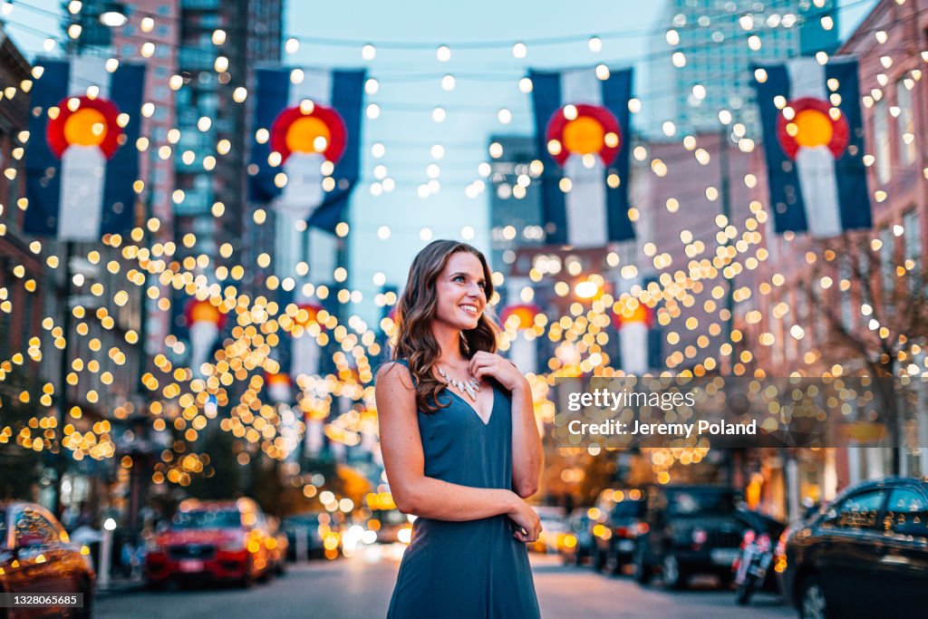Portrait of a Beautiful, Fashionable Young Woman Looking Up and Standing Under Colorado Flags and String Lights on Larimer Square in Denver, Colorado at Dusk