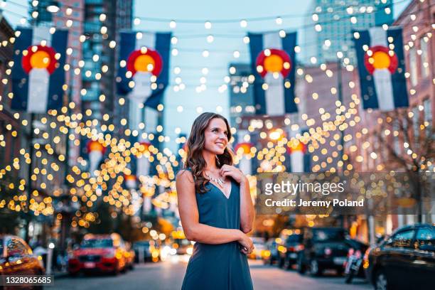 portrait of a beautiful, fashionable young woman looking up and standing under colorado flags and string lights on larimer square in denver, colorado at dusk - denver stock pictures, royalty-free photos & images