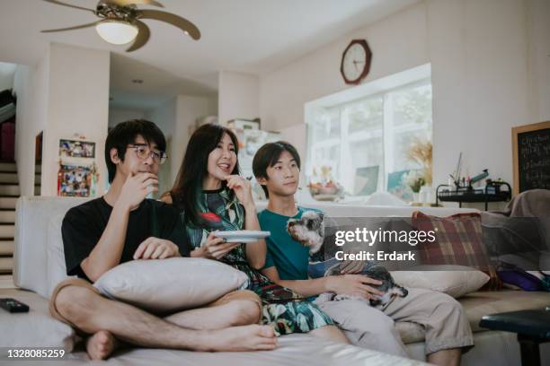 young two asian boys sitting with their mother on sofa in the living room watching tv together on holiday - stock photo - asian watching tv stock pictures, royalty-free photos & images