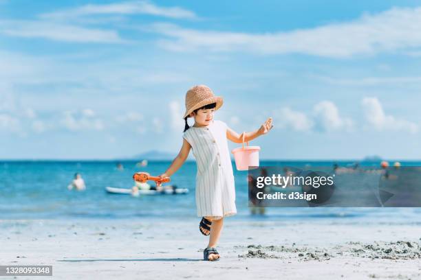 portrait of joyful little asian girl with straw hat smiling happily, holding a sand bucket and shovel having fun at beach on a sunny summer day - beach shovel stockfoto's en -beelden