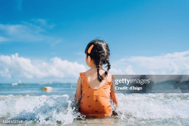 rear view of lovely little asian girl sitting by the seashore at the beach and being splashed by waves. having fun at beach on a sunny summer day - asian girl photos et images de collection