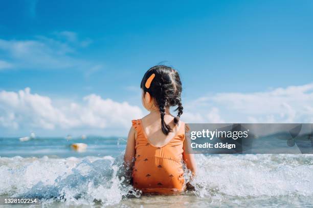 rear view of lovely little asian girl sitting by the seashore at the beach and being splashed by waves. having fun at beach on a sunny summer day - badkläder bildbanksfoton och bilder