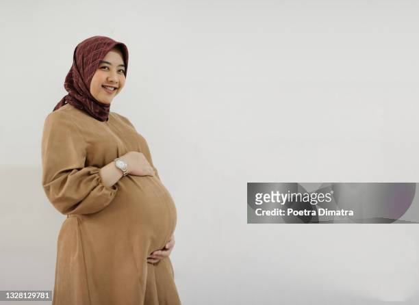 muslim woman holding her pregnant belly and smiling - pregnant muslim stock pictures, royalty-free photos & images