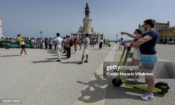 Mask-clad tourists pause with their Link e-scooters to watch demonstrators gathering in Praça do Comercio to protest against current practices by SEF...