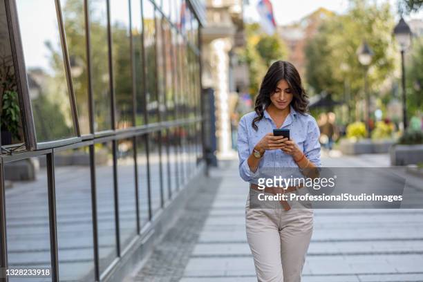 eine junge frau läuft im stadtteil spazieren und smst nachrichten auf ihrem handy. - woman using smartphone with laptop stock-fotos und bilder