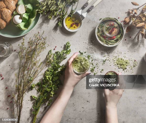 women hands holding homemade flavor herbs salt in jars  on pale concrete kitchen table with dried herbs, bowls, oil, bread and eggs - herbs and spices stock-fotos und bilder