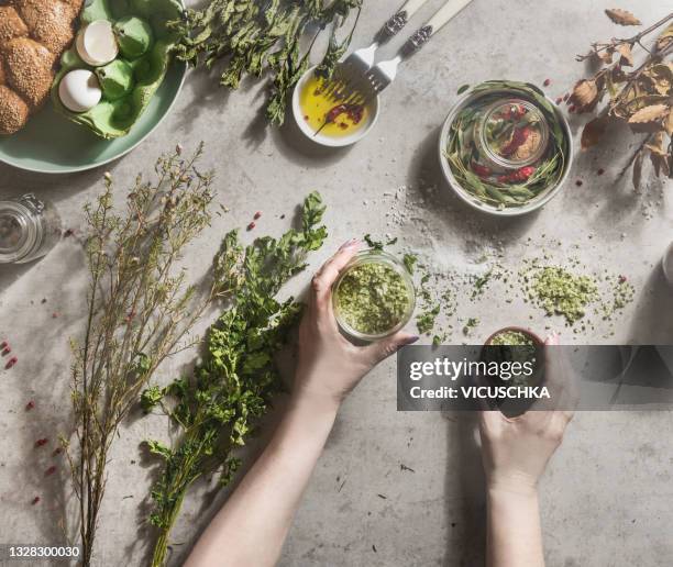 women hands holding homemade flavor herbs salt in jars  on pale concrete kitchen table with dried herbs, bowls, oil, bread and eggs - dried food stock pictures, royalty-free photos & images