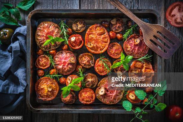 roasted tomatoes cut varied in baking tray and ladle with basil and rosemary on wood - roasted imagens e fotografias de stock
