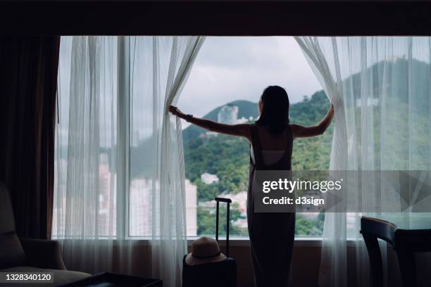 silhouette of young asian female traveller just arriving at the hotel room, opening curtains and looking out to the beautiful scenics through window. with suitcase by her side. travel and vacations concept - curtain hotel stock-fotos und bilder