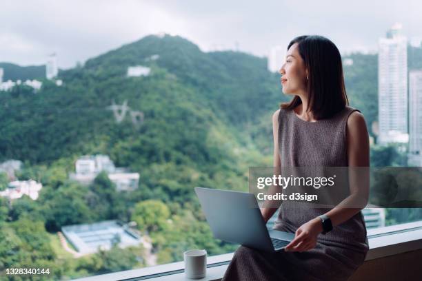 young asian businesswoman looking through window while working on laptop in office, sitting by the window with beautiful view of urban city skyline. female leadership. making business connections with technology - corporate travel stock-fotos und bilder