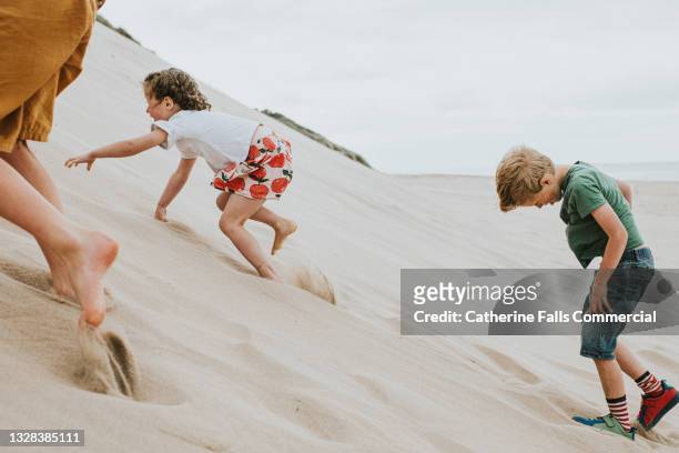 three children clamber up a steep sand dune, which takes a lot of effort as the soft sand gives way under their little feet and hands - beach holiday uk stock pictures, royalty-free photos & images