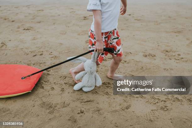 a child walks across a wet beach, dragging a bodyboard behind her while simultaneously clutching her stuffed toy bunny - dirty feet stock-fotos und bilder