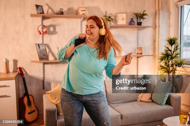 woman holding phone singing and dancing at home - fat woman dancing stockfoto's en -beelden