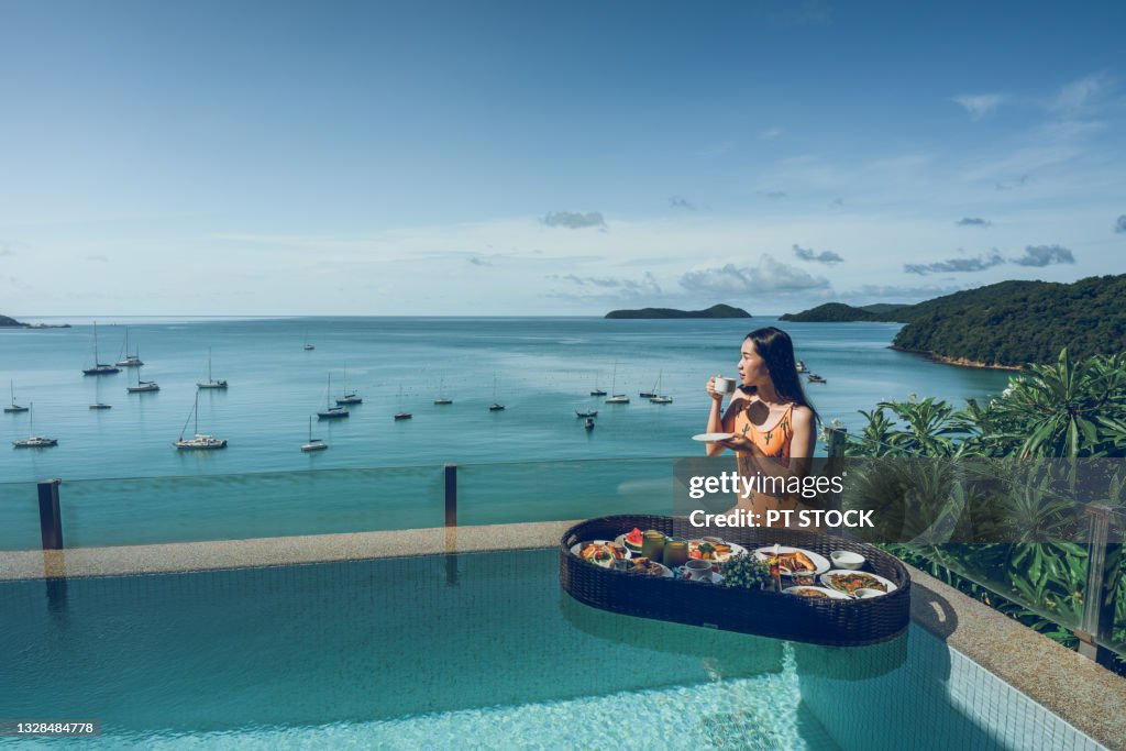 A woman drinking coffee and breakfast floating on the water beside the pool.