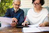 Senior elderly man and woman in eyeglasses examining documents and bills, using calculator considering the amount of rent arrears, checking finances, managing domestic expense,sitting at home terrace