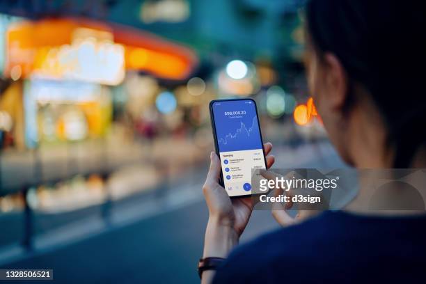over the shoulder view of young asian woman managing online banking with mobile app on smartphone. checking financial data, transferring money, paying bills and checking balances. technology makes life easier. business on the go - financial market photos et images de collection