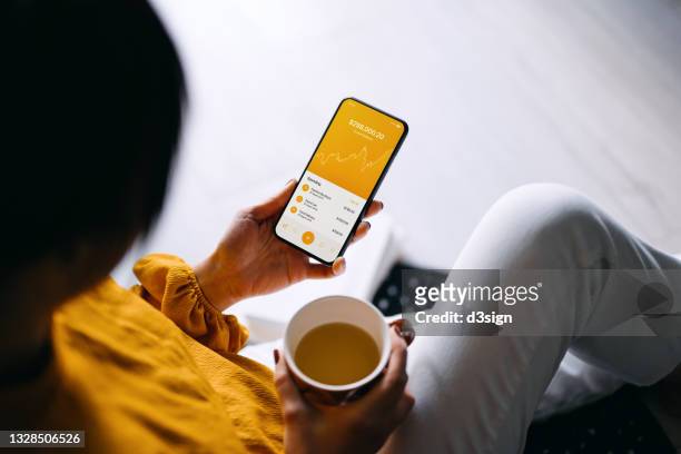over the shoulder view of young asian woman relaxing at cozy home, managing online banking with mobile app on smartphone. checking financial data, transferring money, paying bills and checking balances. technology makes life so much easier - bills payment stock-fotos und bilder