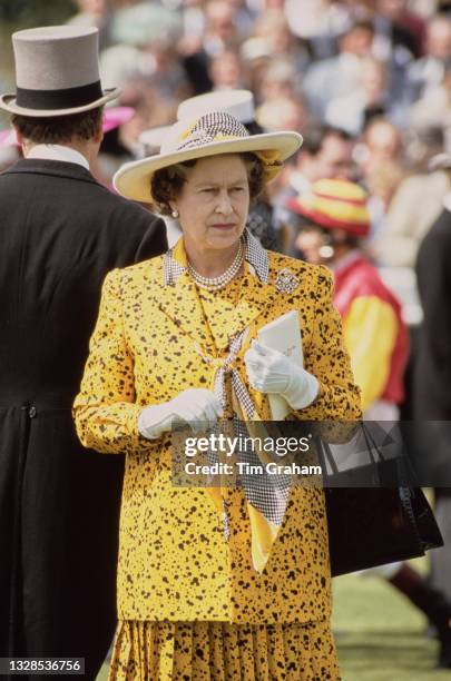 British Royal Queen Elizabeth II wearing a yellow-and-black suit with white gloves and a wide brim hat attends the Derby Day meeting at Epsom Downs...
