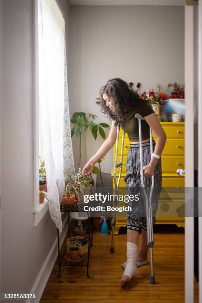 injured millennial woman on crutches spending time with house plants - crutches stockfoto's en -beelden