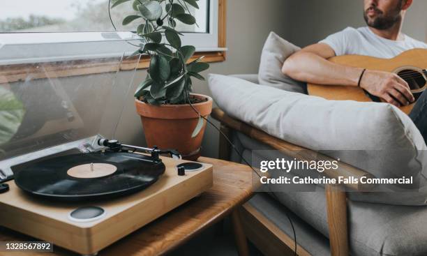 man plays guitar on a comfortable and stylish sofa beside a modern record player. - record player fotografías e imágenes de stock