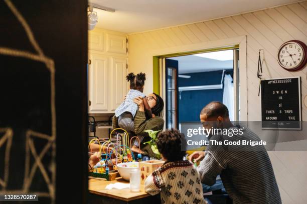 father playing with son in kitchen at home while mother lifts baby in background - stéréotype de la classe moyenne photos et images de collection