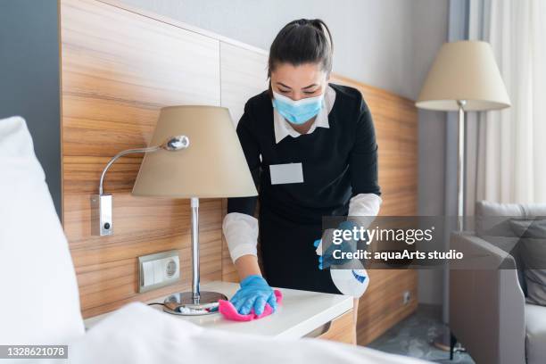 housekeeper with protective face mask cleaning furniture with disinfecting spray in hotel room - hotel housekeeping stock pictures, royalty-free photos & images