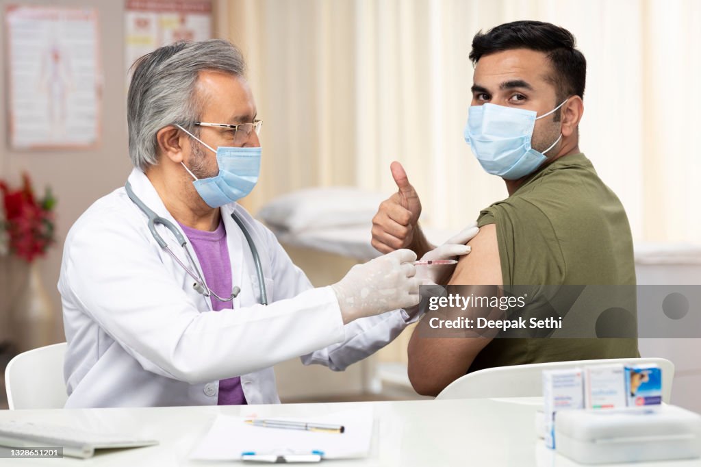 Mature doctor giving COVID-19 or flu antivirus vaccine shot to young men patient's arm at health clinic