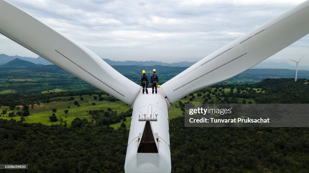 Two Electric engineer wearing Personal protective equipment working on top of wind turbine farm.