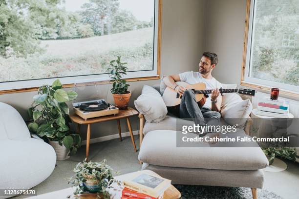 smiling man plays acoustic guitar in a stylish living room. - fabolous musician stock pictures, royalty-free photos & images