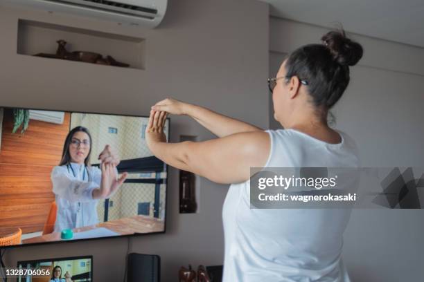 mature woman watching and copying wrist exercises at home, guided by a physical therapist online. - kinesist stockfoto's en -beelden