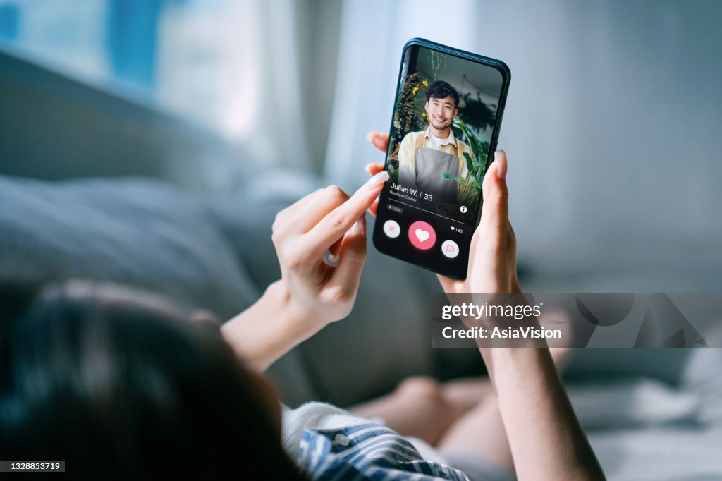 Young Asian woman lying on sofa at home, using an online dating app on smartphone, looking for love on the internet. Social media. Internet dating. Couple relationship. Love concept