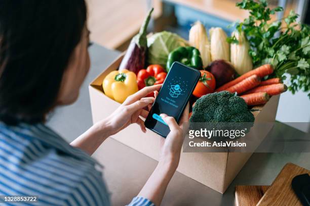over the shoulder view of young asian woman doing home delivery grocery shopping online with mobile app device on smartphone at home, with a box of colourful and fresh organic vegetables and fruits on the table - delivering imagens e fotografias de stock