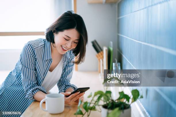 beautiful smiling young asian woman using smartphone and having a cup of coffee in the kitchen at cozy home. lifestyle and technology concept - candid forum 個照片及圖片檔