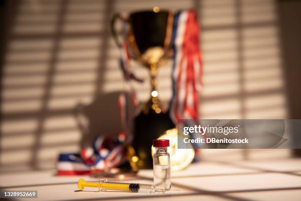 champion's cup and gold medals hanging with the light coming through the window, leaving the shadow of a blind at sunset, next to syringe with doping substance. - doping ストックフォトと画像