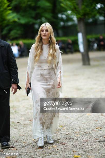 Ioanna Gika wears a white with gold embroidered flower long flowing dress with puffy / cut-out sleeves, a black shiny leather clutch, gold rings,...