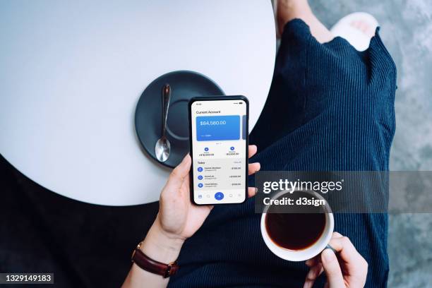 overhead view of young woman managing online banking with mobile app on smartphone while drinking a cup of coffee at sidewalk cafe. tracking and planning spending. transferring money, paying bills, checking balances. technology makes life much easier - bills payment stock-fotos und bilder