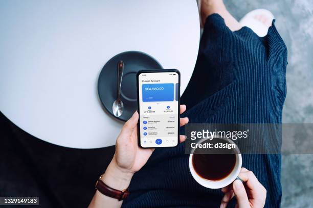 overhead view of young woman managing online banking with mobile app on smartphone while drinking a cup of coffee at sidewalk cafe. tracking and planning spending. transferring money, paying bills, checking balances. technology makes life much easier - coffee chat stockfoto's en -beelden