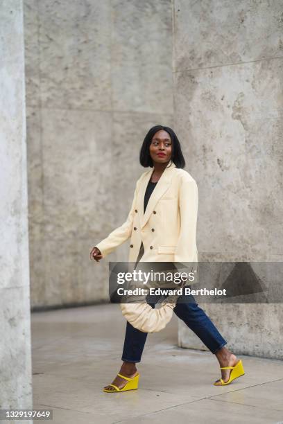 Carrole Sagba @linaose wears a beige oversized blazer jacket, a black top, blue denim jeans pants, a beige leather bag, a golden ring, yellow...