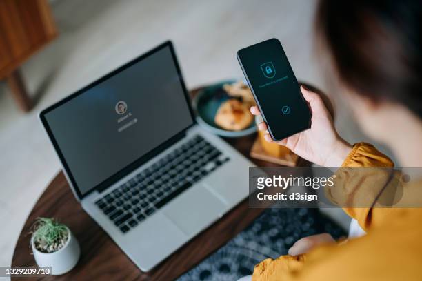 young asian woman logging in to her laptop and holding smartphone on hand with a security key lock icon on the screen, sitting in the living room at cozy home. privacy protection, internet and mobile security concept - privat stock-fotos und bilder