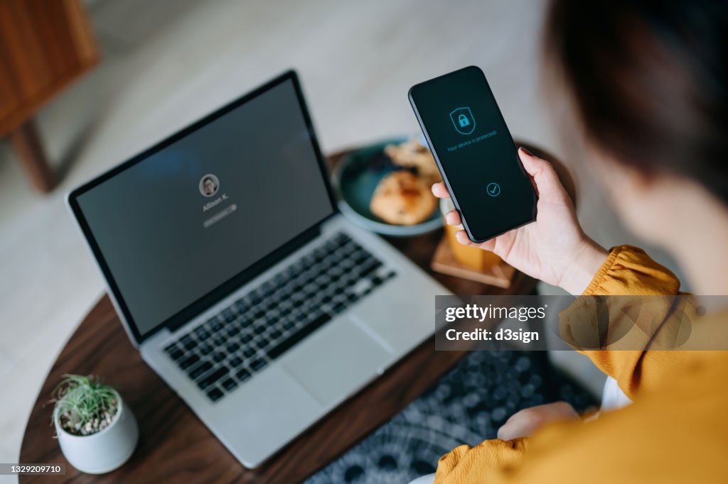 Young Asian woman logging in to her laptop and holding smartphone on hand with a security key lock icon on the screen, sitting in the living room at cozy home. Privacy protection, internet and mobile security concept