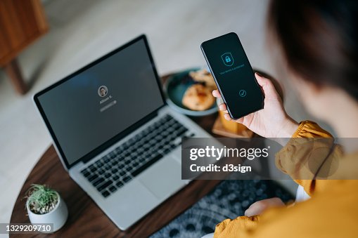 Young Asian woman logging in to her laptop and holding smartphone on hand with a security key lock icon on the screen, sitting in the living room at cozy home. Privacy protection, internet and mobile security concept Young Asian woman logging in to her laptop and holding smartphone on hand with a security key lock icon on the screen, sitting in the living room at cozy home. Privacy protection, internet and mobile security concept