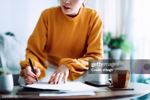 cropped shot of young asian woman holding a pen and signing paperwork in the living room at home. deal concept - deal signing stockfoto's en -beelden