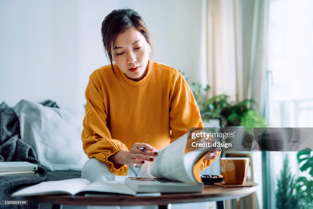 Focused young Asian woman reading book and making notes at home, concentrates on her studies. Further education concept