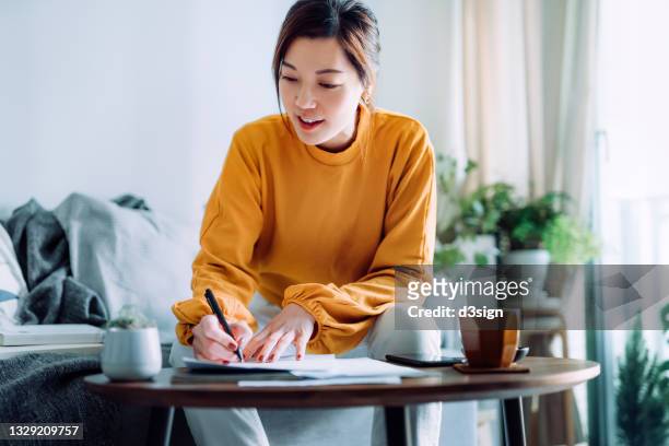 young asian woman holding a pen and signing paperwork in the living room at home. deal concept - asian finance photos et images de collection