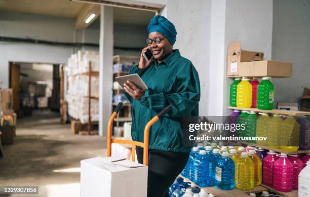 afro female warehouse worker using a hand truck and talking on a smart phone in distribution warehouse - country market stock pictures, royalty-free photos & images